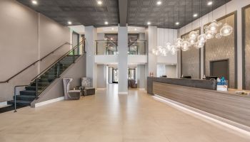 The image shows a modern hotel lobby with a staircase, a reception desk, stylish lighting fixtures, and seating areas in a spacious and elegant setting.