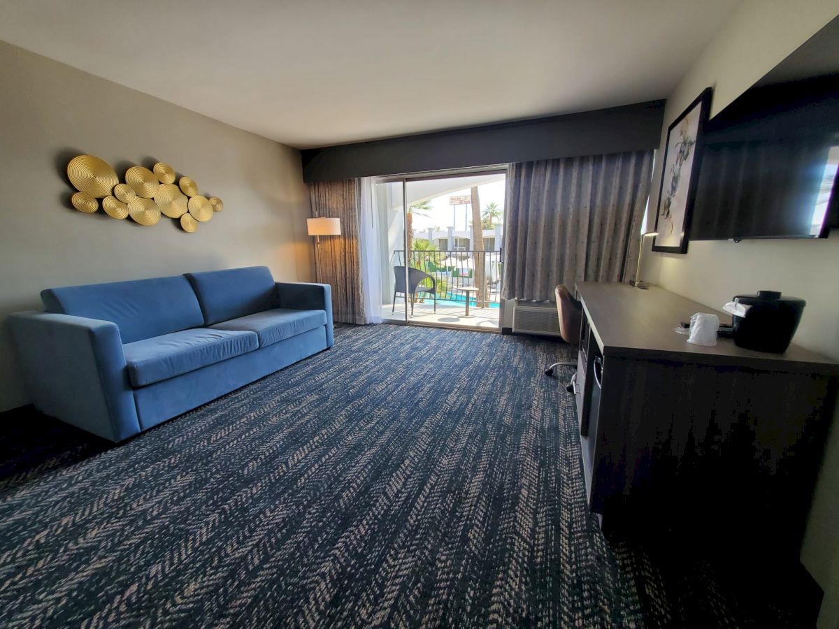 A modern hotel room with a blue sofa, desk, wall art, TV, and large window with balcony offering a view of an outdoor pool area.