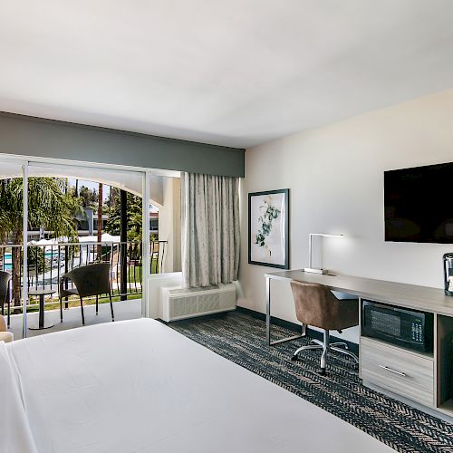 A hotel room with a bed, TV, desk, coffee maker, and a sliding door leading to a balcony with outdoor seating and a pool view ending the sentence.