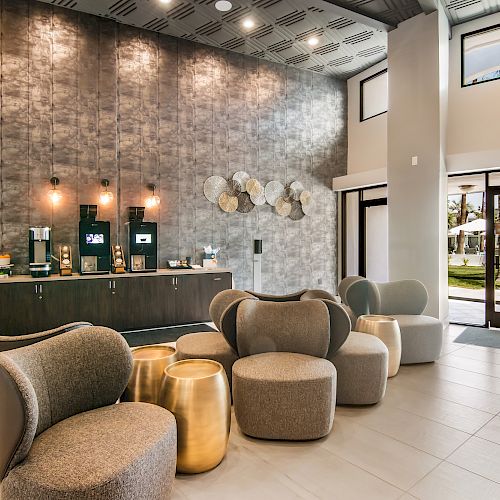 A modern lobby with stylish seating, golden side tables, and a coffee station; there's a view of an outdoor area through the glass doors.