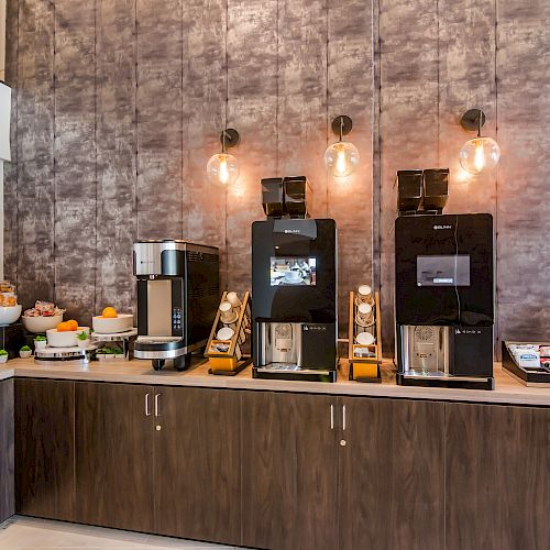 The image shows a modern coffee station with three coffee machines, a boiling water dispenser, and an assortment of cups and condiments on a counter.