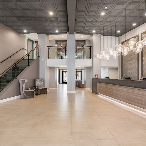 A modern hotel lobby with minimalist decor features a seating area, a grand staircase, a reception desk, and stylish hanging lights, elegantly arranged.