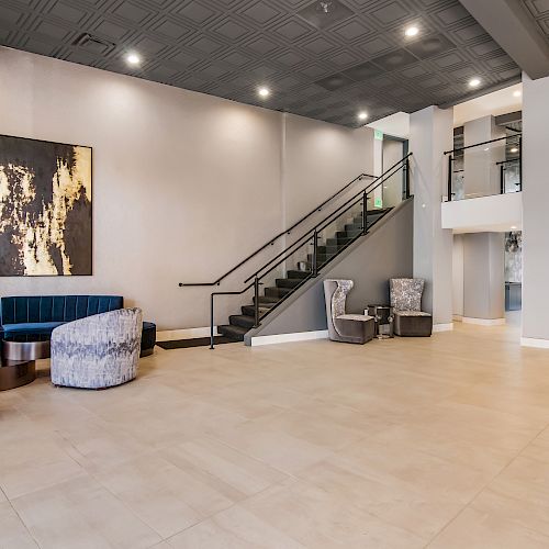 A modern lobby with seating area, artwork on the wall, stairs leading to the upper level, light tiles, and ample space for movement.