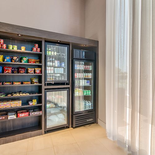 The image shows a snack and beverage station with shelves of packaged snacks and two refrigerators containing drinks, located in a modern, well-lit room.