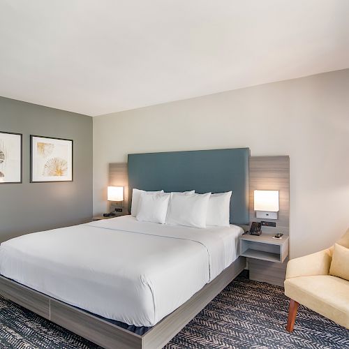 A modern hotel room with a large bed, two bedside lamps, framed artwork, a beige armchair, and gray carpeting.