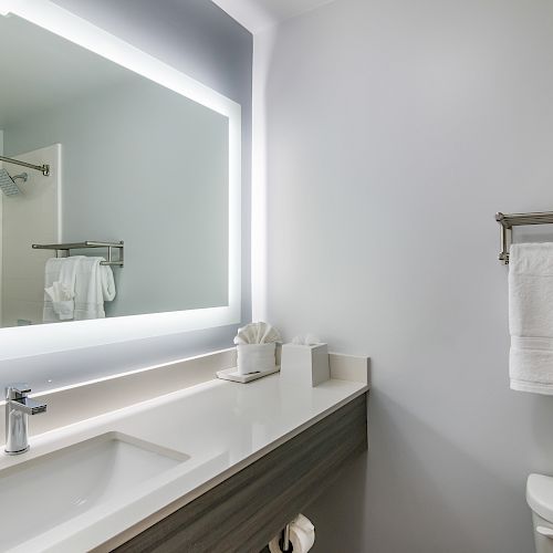 A modern, clean bathroom with a large, illuminated mirror, sink, towel rack with neatly folded towels, and a toilet in a minimalist style.