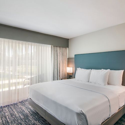 A modern hotel room features a neatly made king-size bed, bedside tables with lamps, a telephone, large window, and light gray curtains.