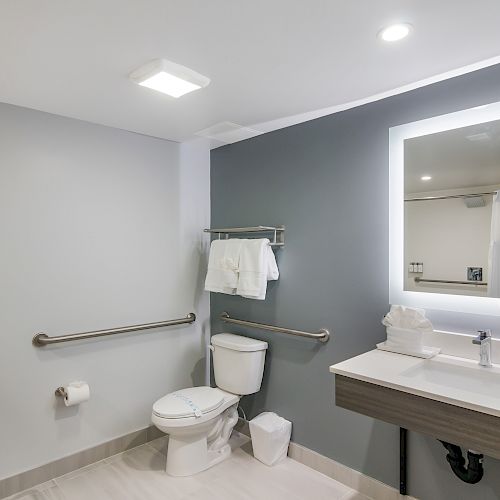 A modern bathroom with a toilet, grab bars, a sink with a lit mirror, a towel rack with towels, and a shower chair against a light gray wall.