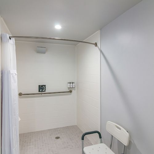 The image shows an accessible shower with a handheld shower head, a grab bar, a shower chair, and soap dispensers on the wall.