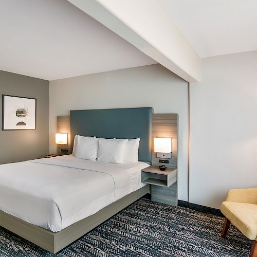 A modern hotel room features a neatly made bed, bedside tables with lamps, wall art, and an upholstered chair in a well-lit setting.