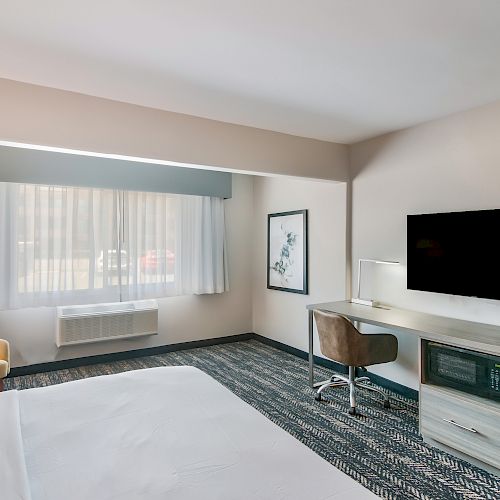 The image shows a modern hotel room with a bed, a TV, a desk with a chair, a mini fridge, and a beige chair by the window with curtains.