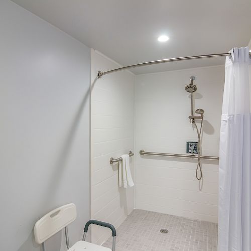 This image shows an accessible shower equipped with a shower chair, grab bar, handheld showerhead, and a curtain partially drawn.