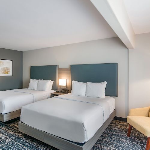 The image shows a modern hotel room with two neatly made double beds, blue padded headboards, framed art on the wall, a soft chair, and ample lighting.