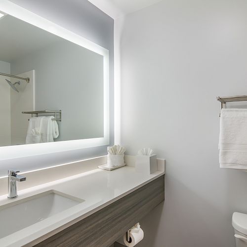 This image shows a modern bathroom with a large backlit mirror, sink, towel rack with folded towels, and a toilet.