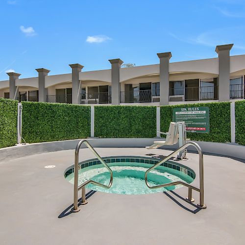 The image shows an outdoor Jacuzzi surrounded by a paved area and greenery, with a building in the background and a clear blue sky.