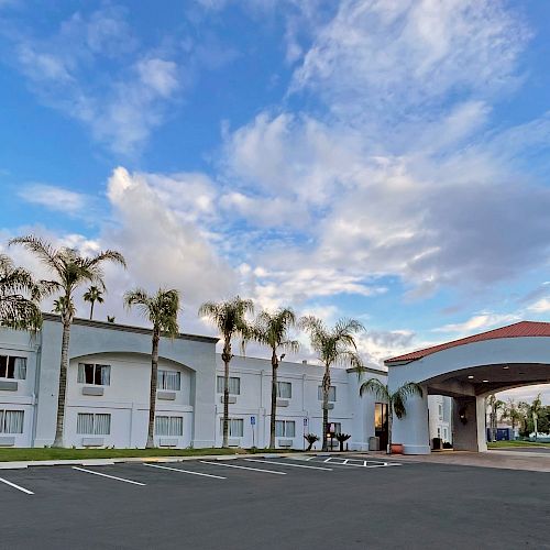 The image shows a two-story white building, likely a hotel, with an arched entrance, palm trees, and an adjacent parking lot under a partly cloudy sky.