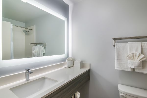 A modern bathroom featuring a large illuminated mirror, a sink with a sleek faucet, towels folded neatly, and a shower area in the background.