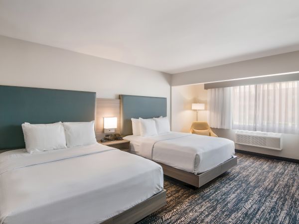 A hotel room with two neatly made double beds, nightstand, lamp, armchair, large window with curtains, carpeted floor, and modern decor.