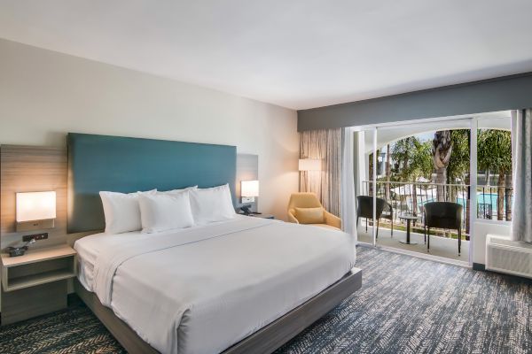 A modern hotel room with a large bed, two bedside tables, a cozy chair, and a balcony overlooking a pool area, creating a relaxing atmosphere.