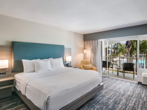 A modern hotel room with a large bed, two bedside tables, a cozy chair, and a balcony overlooking a pool area, creating a relaxing atmosphere.