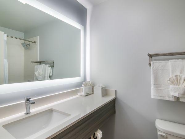 This image shows a modern bathroom with a large, illuminated mirror, sink, towel rack with folded towels, tissue box, and a toilet.