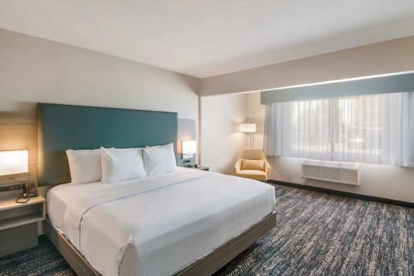 A hotel room featuring a large bed, bedside tables with lamps, a yellow armchair by the window, and patterned carpet flooring, all in neutral tones.