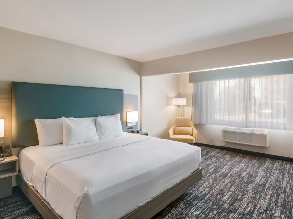 A hotel room featuring a large bed, bedside tables with lamps, a yellow armchair by the window, and patterned carpet flooring, all in neutral tones.