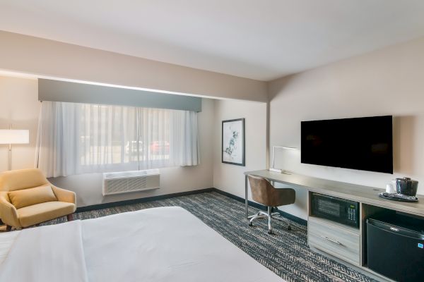 A modern hotel room with a bed, TV, armchair, desk, lamp, microwave, mini-fridge, and coffee maker, decorated in neutral tones, and a window allowing natural light.