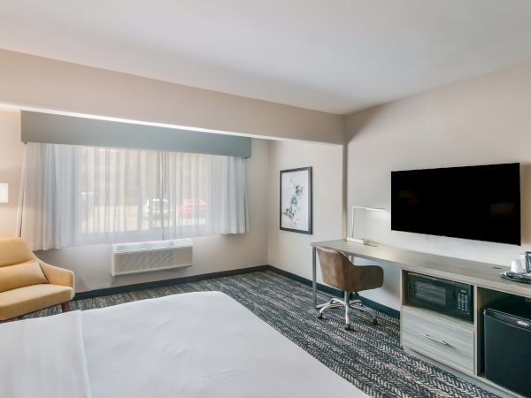A modern hotel room with a bed, TV, armchair, desk, lamp, microwave, mini-fridge, and coffee maker, decorated in neutral tones, and a window allowing natural light.