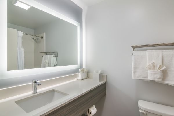 The image shows a modern bathroom with a large illuminated mirror, a sink, neatly folded towels, and a toilet, creating a clean ambiance.