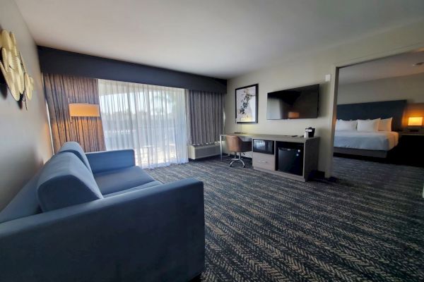 This image shows a modern hotel room with a sofa, flat-screen TV, desk, mini-fridge, and a bed in an adjoining room, plus natural light from large windows.