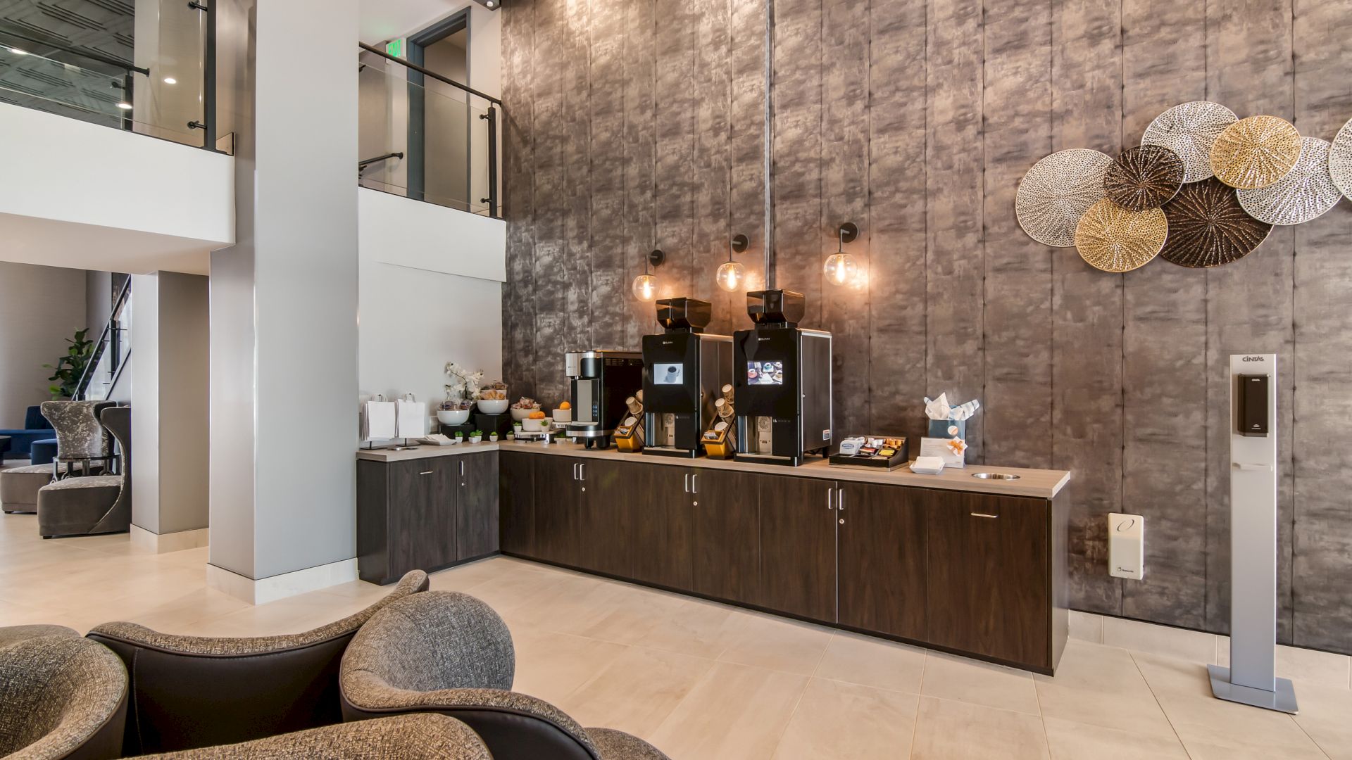 The image shows a modern coffee station in a hotel or office lobby, featuring coffee machines, pastries, and a stylish seating area with cozy chairs.