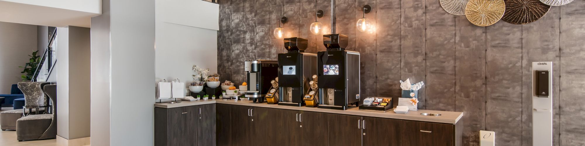 The image shows a modern coffee station in a hotel or office lobby, featuring coffee machines, pastries, and a stylish seating area with cozy chairs.