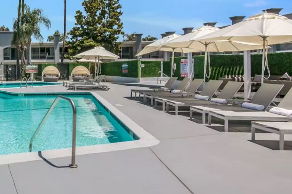 The image shows a modern outdoor swimming pool area with lounge chairs, umbrellas, and cabanas in a sunny, resort-like setting.