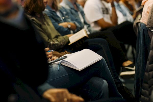 A group of people sitting close together, with some holding notebooks and pens, suggesting they might be attending a class or seminar.