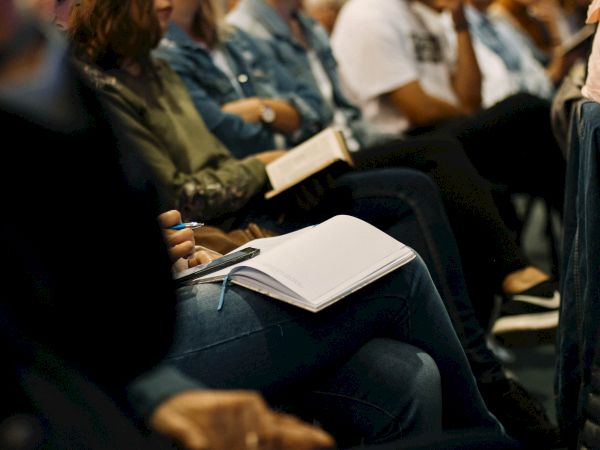 A group of people sitting close together, with some holding notebooks and pens, suggesting they might be attending a class or seminar.