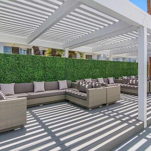 The image shows an outdoor seating area with a white pergola, light-colored wicker furniture, cushions, and a backdrop of greenery, casting striped shadows.