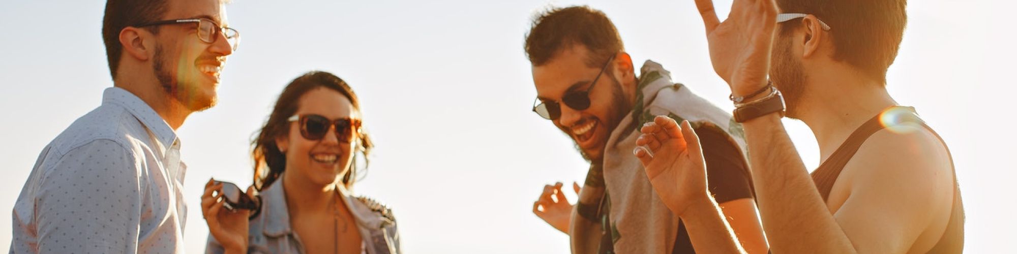 A group of four people are outdoors, enjoying each other's company, laughing and talking with a sunny backdrop in the background.