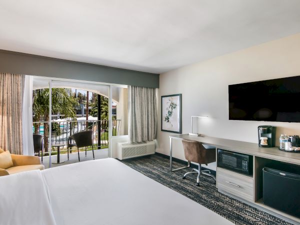A hotel room with a bed, desk, TV, coffee maker, and mini-fridge. There's a balcony with chairs overlooking a pool area outside.