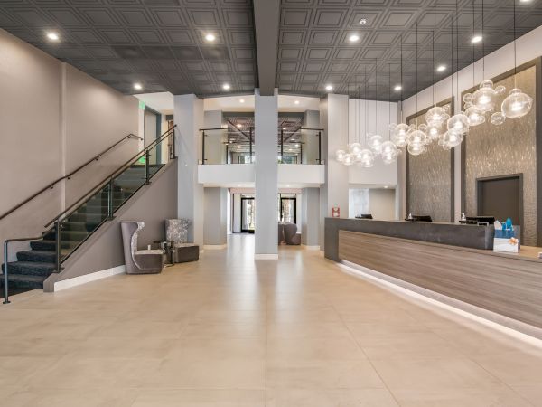 The image shows a modern, spacious hotel lobby with a reception counter, seating area, glass staircase, and contemporary lighting fixtures.
