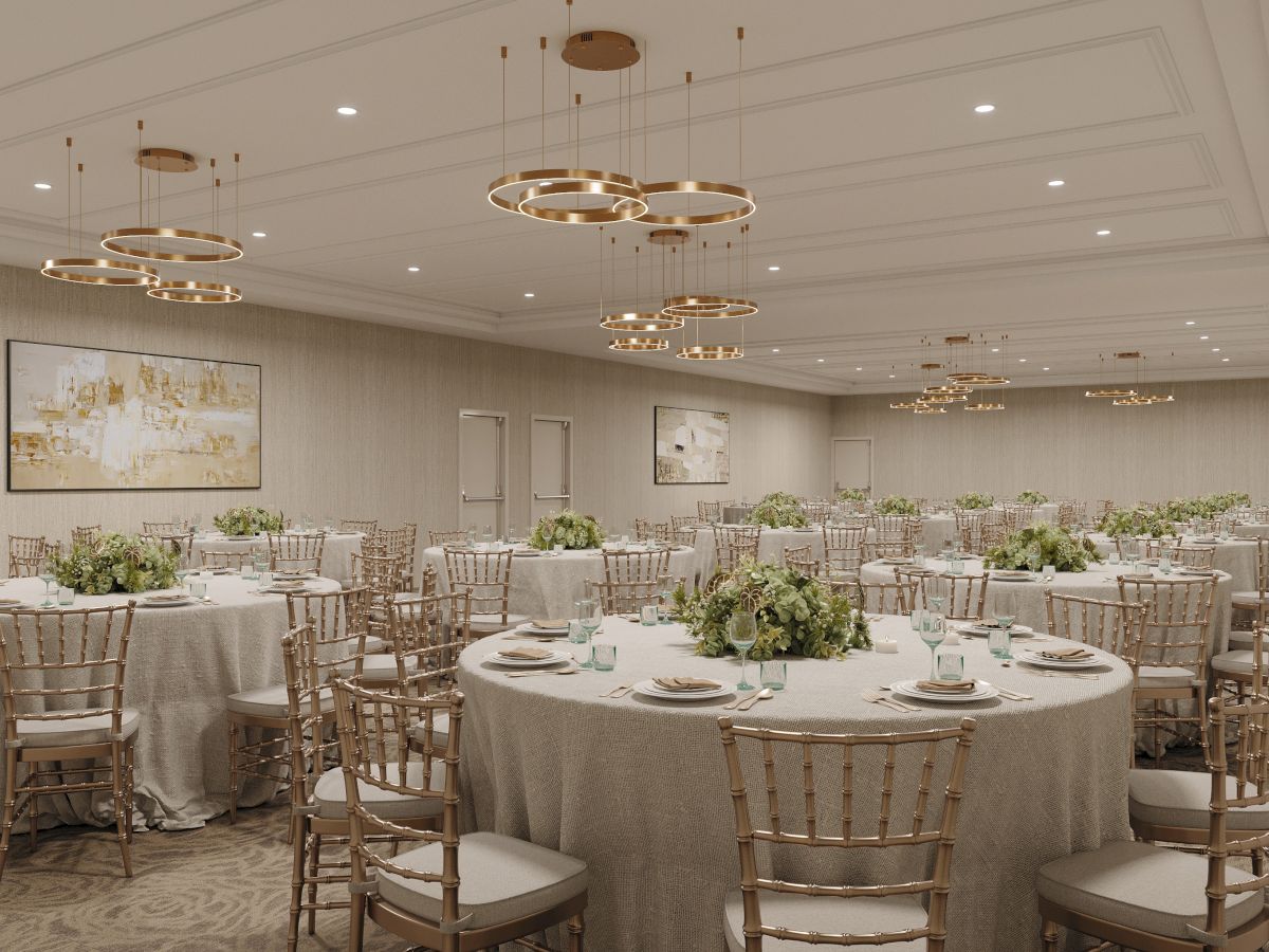 An upscale banquet hall with round tables, gold chiavari chairs, floral centerpieces, and glowing ring chandeliers overhead.