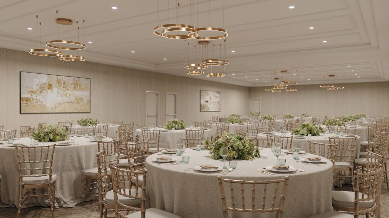 An upscale banquet hall with round tables, gold chiavari chairs, floral centerpieces, and glowing ring chandeliers overhead.
