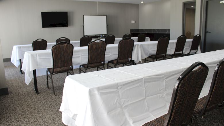 The image shows a conference room setup with rows of chairs and tables covered in white cloths, a TV, and a whiteboard at the front.