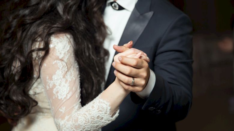 A couple dressed formally, holding hands and dancing. The woman is in a lace dress, and the man is in a black suit with a bow tie, both partially visible.