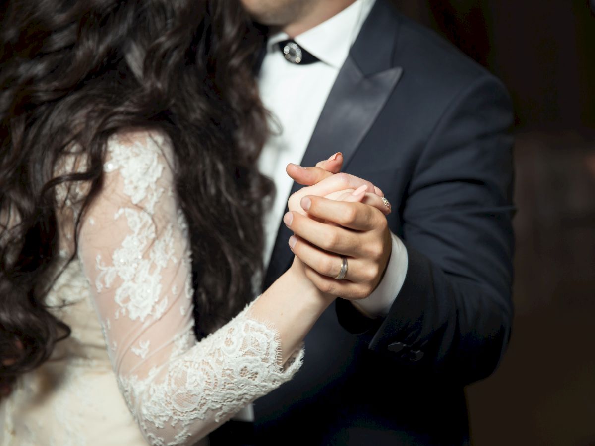 A couple is dancing closely, with the woman's hand in the man's, and the woman wearing a lace dress and the man in a suit and bow tie.