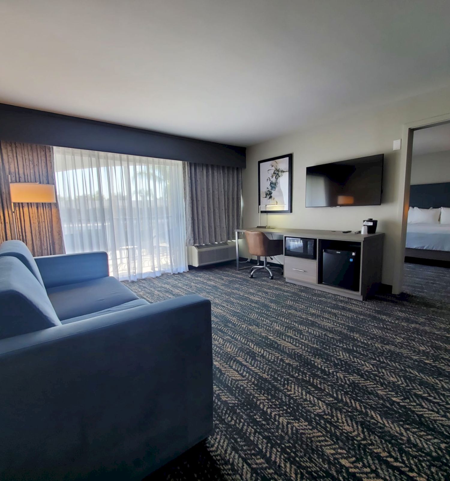 A modern hotel room with a blue sofa, desk, TV, mini-fridge, and adjoining bedroom with a king-size bed. Large window with curtains.