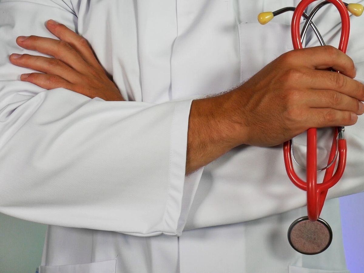 A person in a white lab coat is holding a red stethoscope and has their arms crossed.