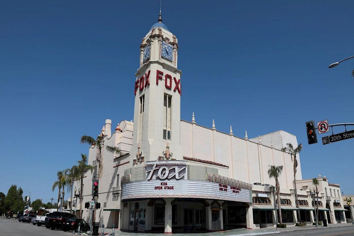 The image shows a historic theater named "Fox" with an art deco design and a tall clock tower, located on a street corner.