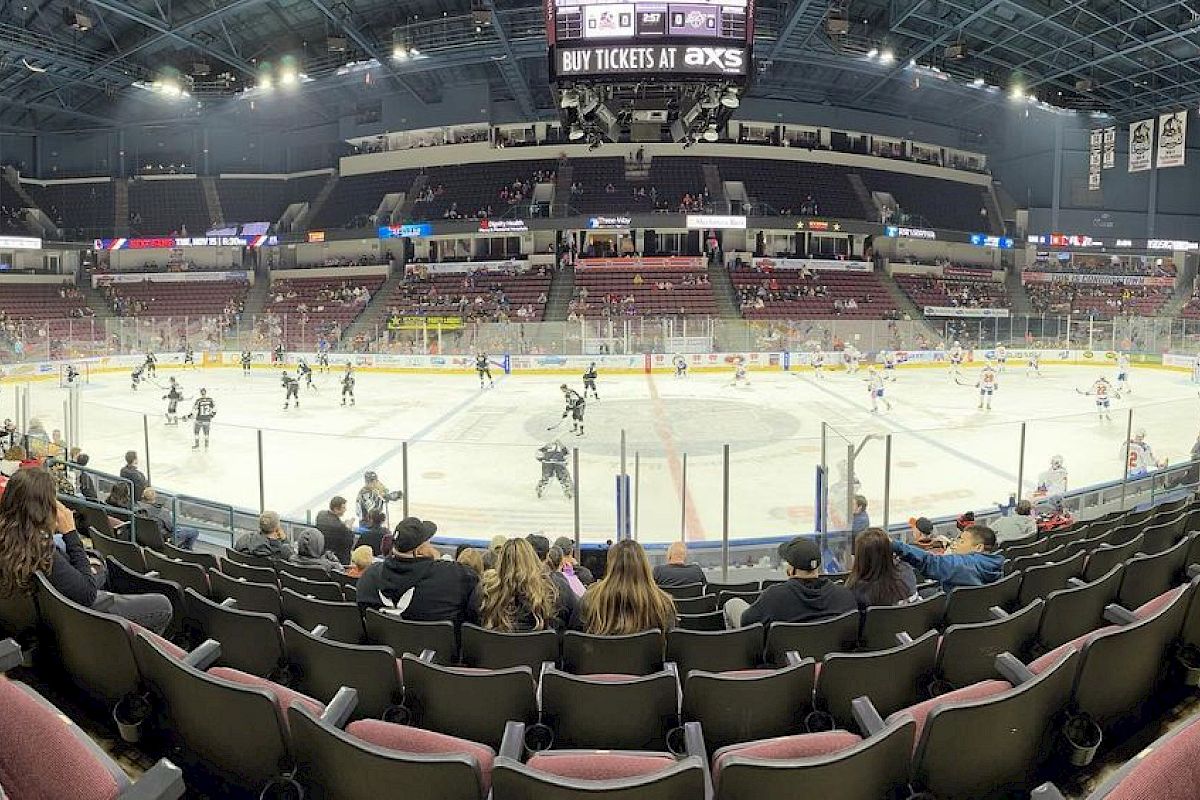 People are watching a hockey game in an indoor arena with players on the ice and a scoreboard above.