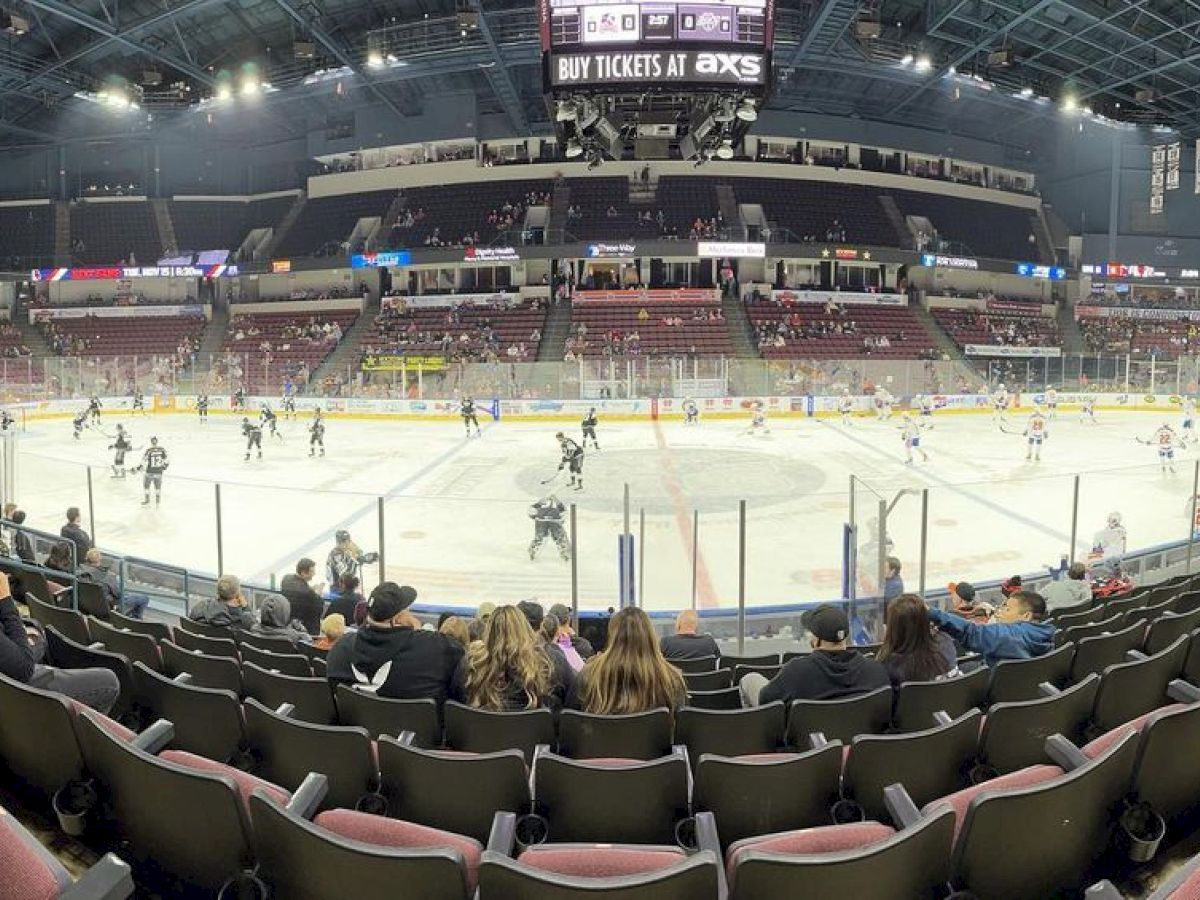People are watching a hockey game in an indoor arena with players on the ice and a scoreboard above.